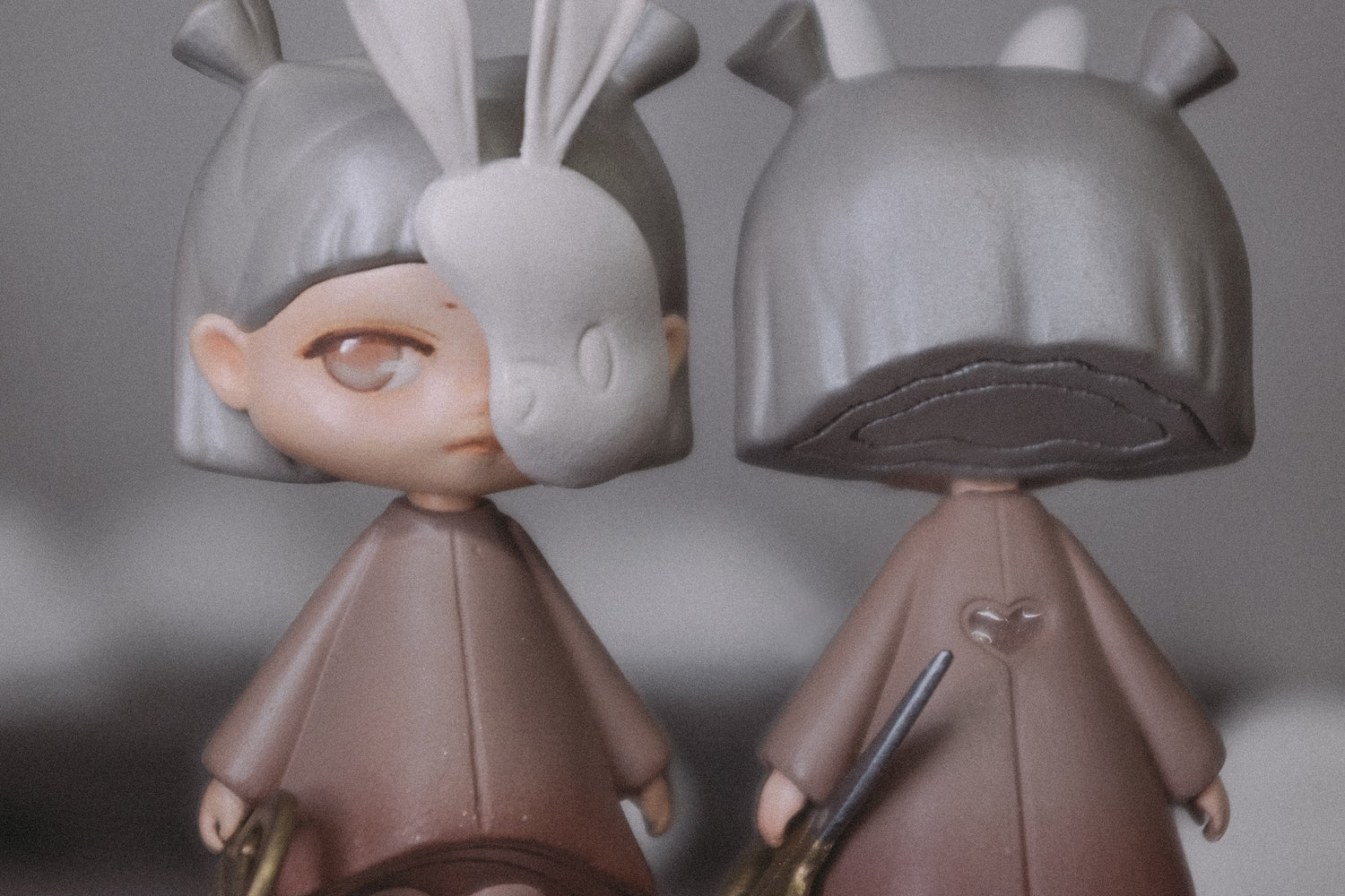 Two stylized doll figurines with oversized bunny-ear masks and long hair, shown standing side by side against a soft gray blurred background.
