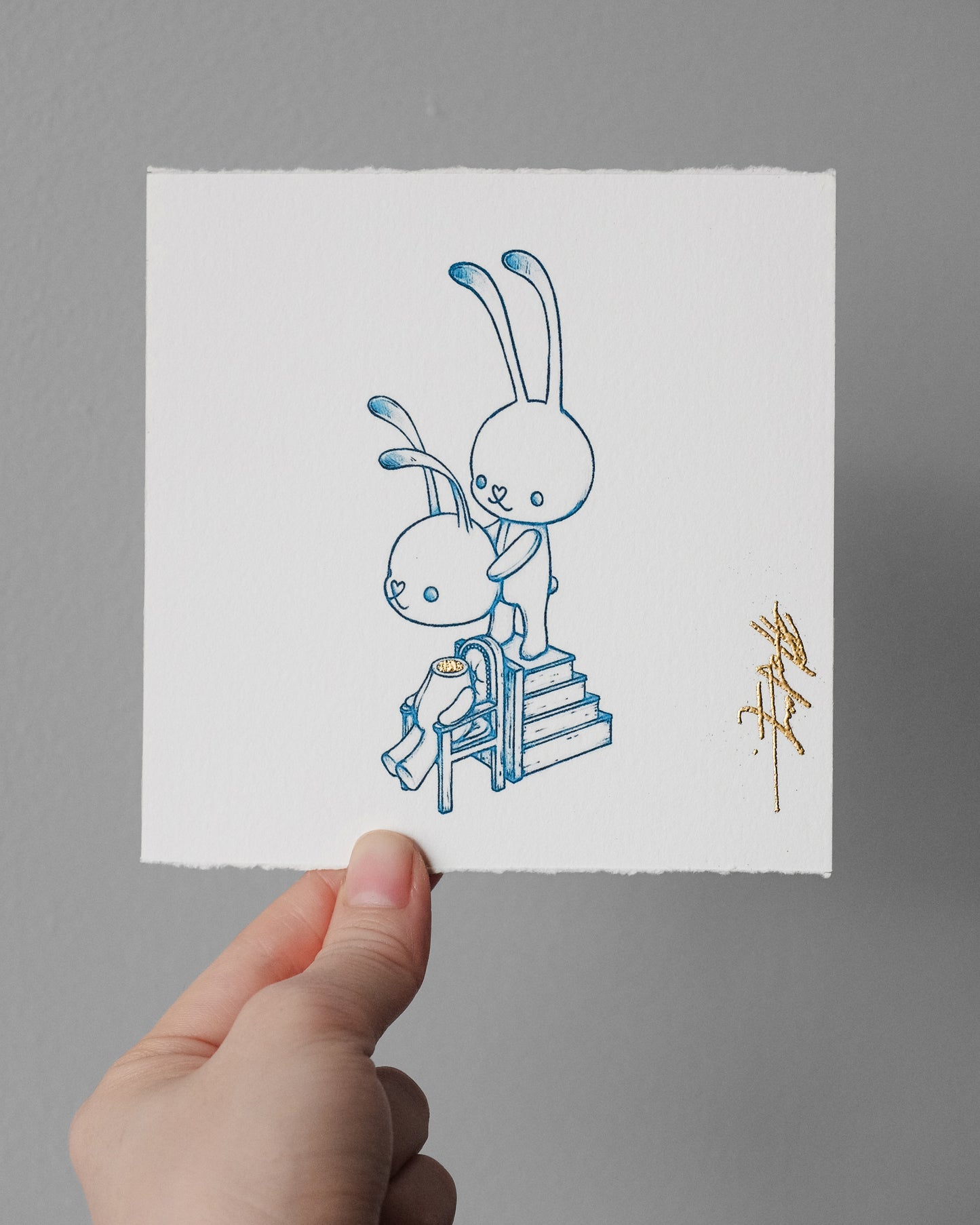 A hand holds a square white art print featuring a minimalist blue line drawing of two rabbits and a small gold signature, against a plain gray wall.