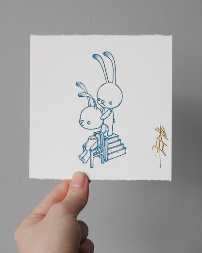 A hand holds a square white art print featuring a minimalist blue line drawing of two rabbits and a small gold signature, against a plain gray wall.
