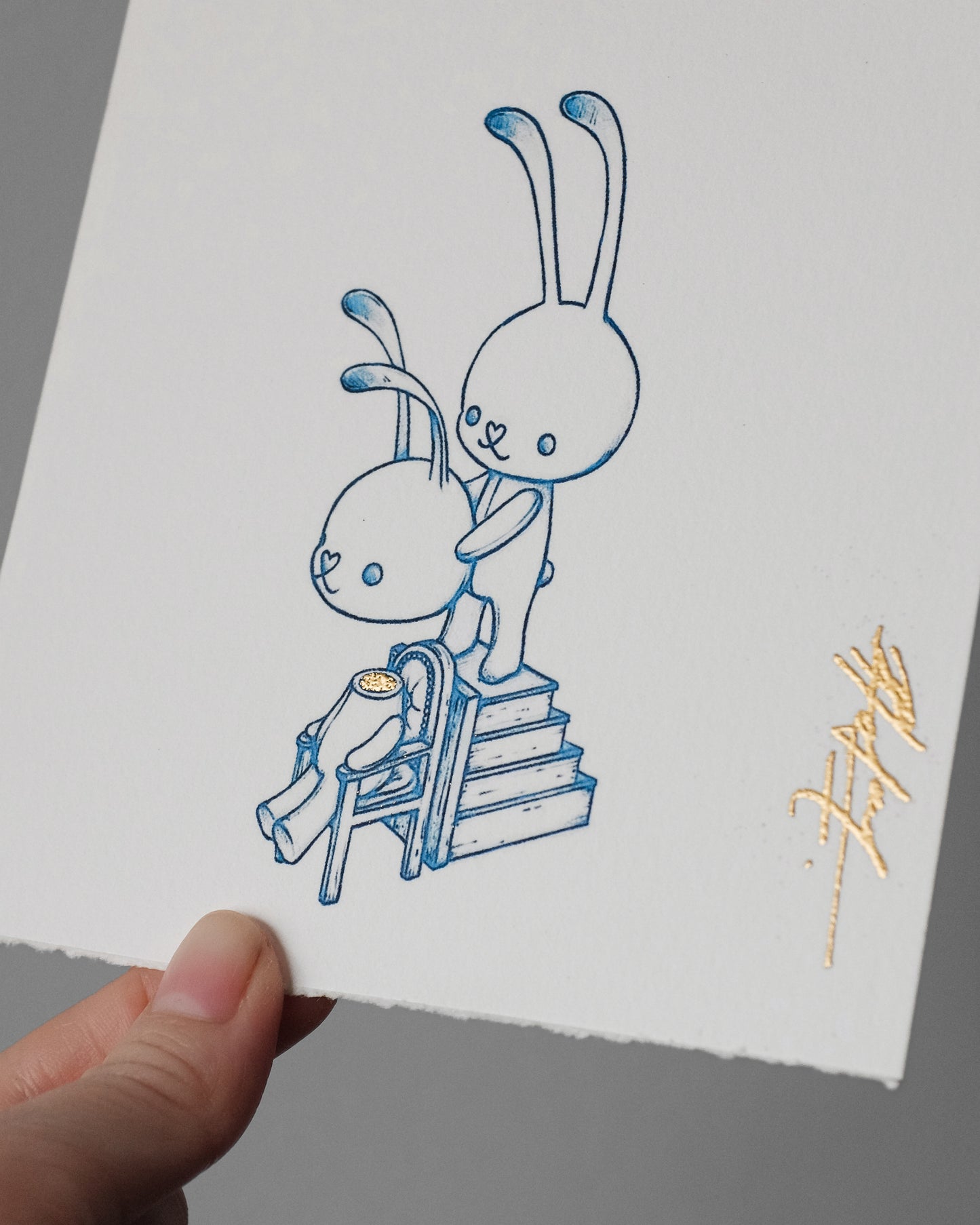 A hand holds a white sheet of textured paper featuring a blue line-art illustration of two cartoon rabbits, one standing on steps behind the other, with a small gold signature in the lower right corner.