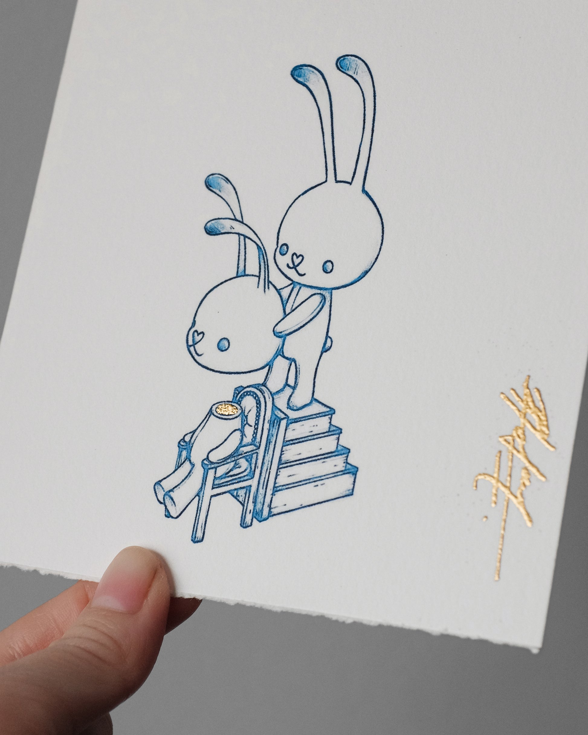 A hand holds a white sheet of textured paper featuring a blue line-art illustration of two cartoon rabbits, one standing on steps behind the other, with a small gold signature in the lower right corner.