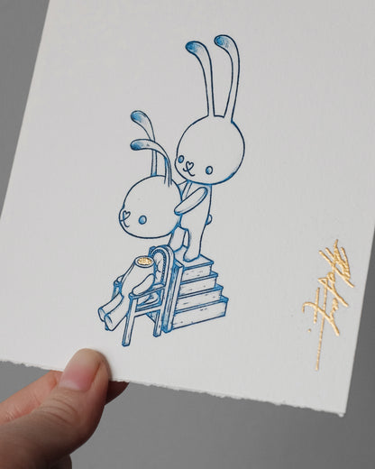 A hand holds a white sheet of textured paper featuring a blue line-art illustration of two cartoon rabbits, one standing on steps behind the other, with a small gold signature in the lower right corner.