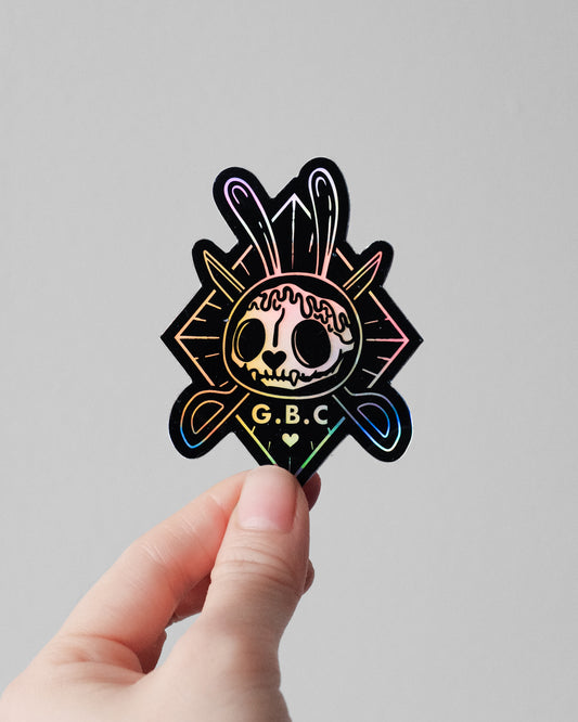 A hand holding a die-cut sticker featuring a stylized skull with a rainbow holographic foil finish, set against a plain light gray background.