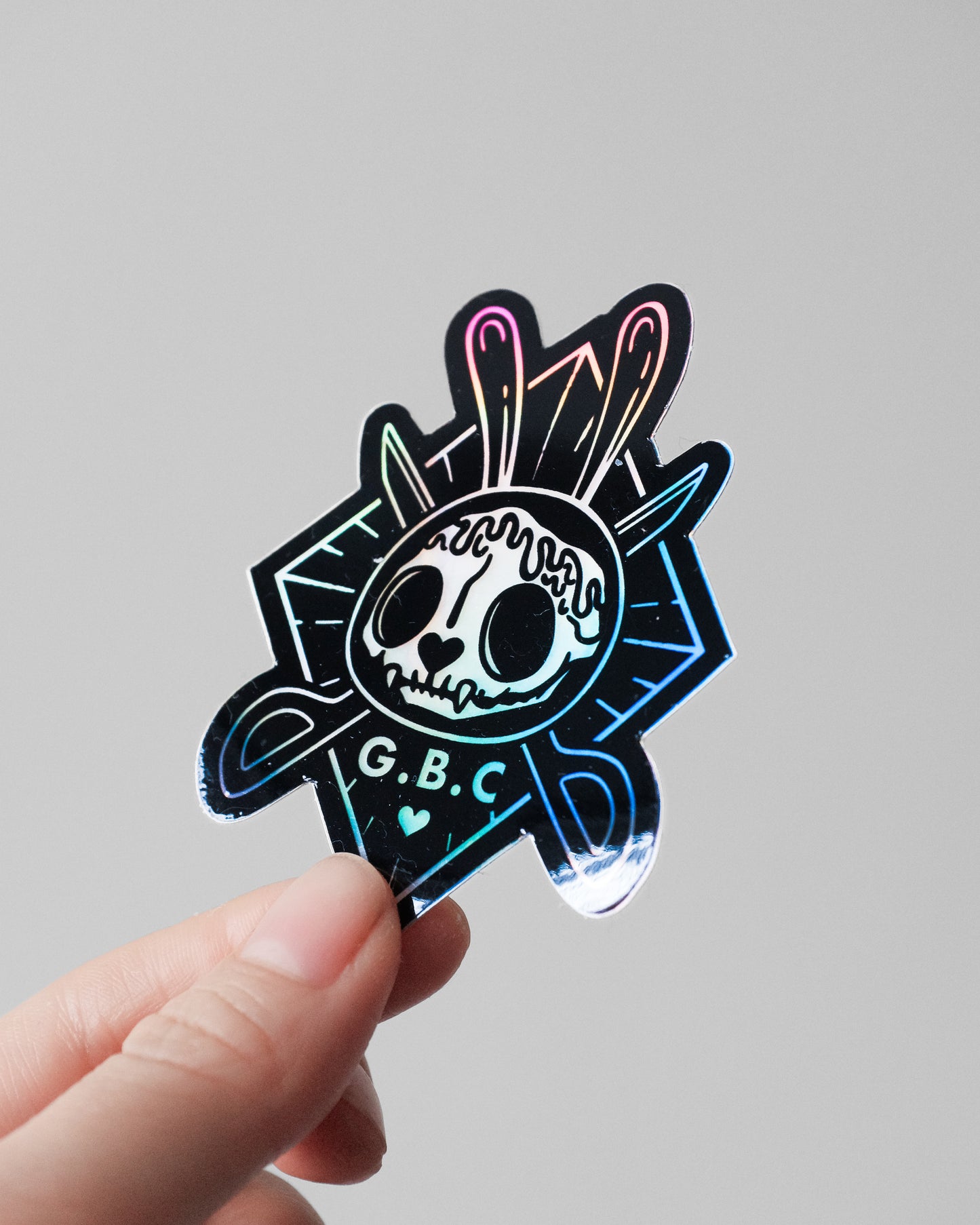 A hand holding a die-cut holographic sticker featuring a stylized skull with bunny ears and the text 'G.B.