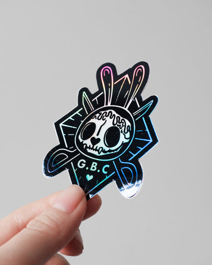 A hand holding a die-cut holographic sticker featuring a stylized skull with bunny ears and the text 'G.B.