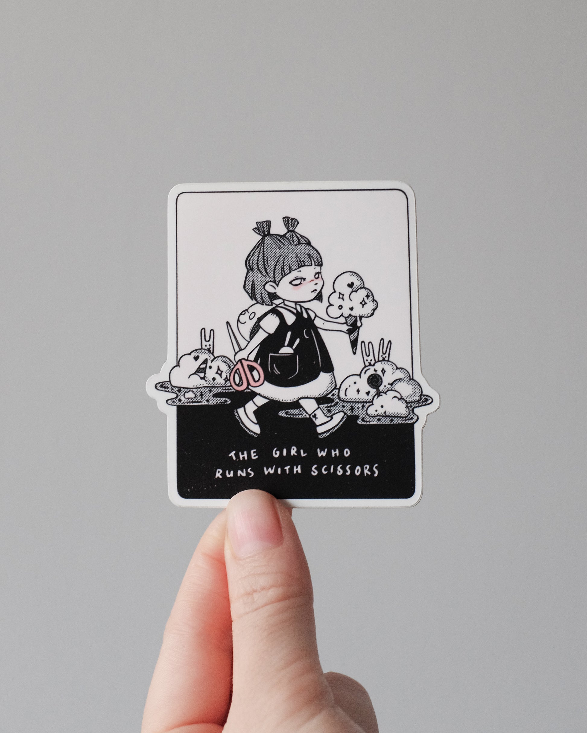 A hand holds a rectangular sticker with rounded corners against a plain light gray background. The sticker features a black and white illustration of a girl with pigtails holding scissors and food, with the text 'THE GIRL WHO RUNS WITH SCISSORS' at the bottom.